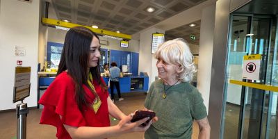 Operatrice di Poste Italiane mostra l'utilizzo dell'app Poste a una cliente anziana all'interno di un ufficio postale, promuovendo l'inclusione digitale tra le persone over 80.