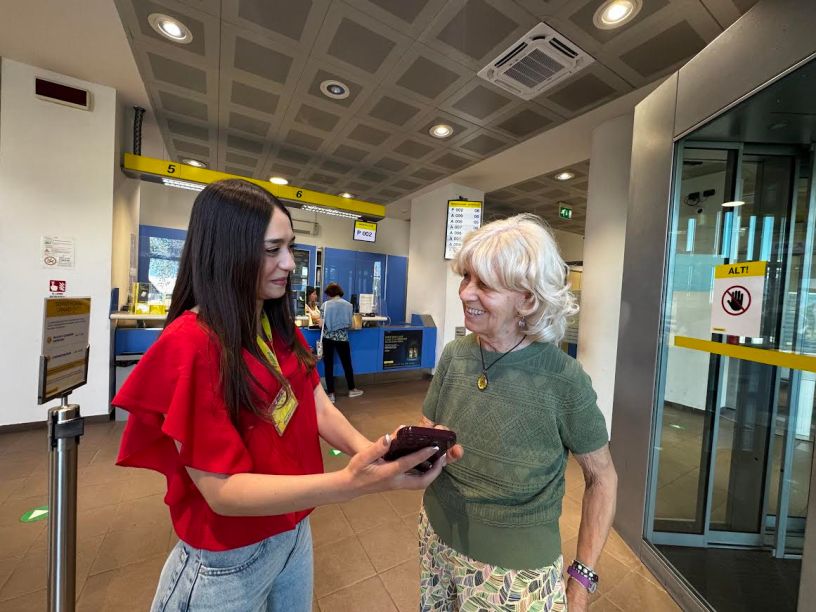 Operatrice di Poste Italiane mostra l'utilizzo dell'app Poste a una cliente anziana all'interno di un ufficio postale, promuovendo l'inclusione digitale tra le persone over 80.