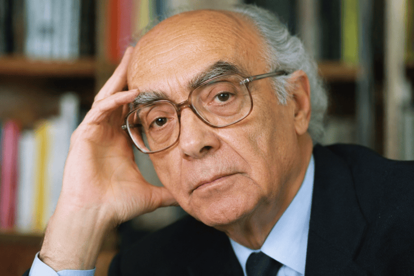 Ritratto fotografico realistico di José Saramago nel suo studio, circondato da libri, mentre riflette con sguardo intenso e pensieroso.