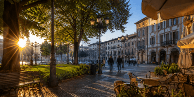 Piazza italiana illuminata dal sole del mattino, con persone che passeggiano tra alberi, caffè all'aperto e palazzi storici in un'atmosfera serena e accogliente.