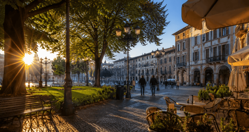 Piazza italiana illuminata dal sole del mattino, con persone che passeggiano tra alberi, caffè all'aperto e palazzi storici in un'atmosfera serena e accogliente.