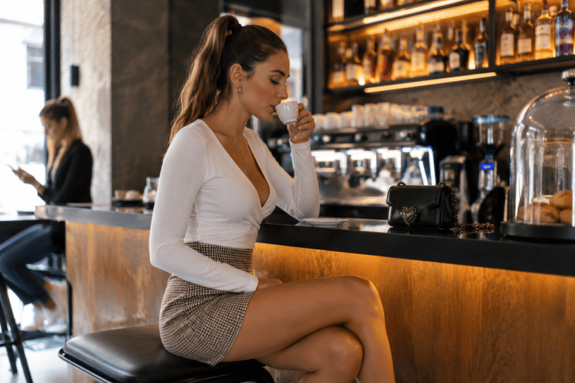 Giovane donna con capelli raccolti in una coda di cavallo mentre sorseggia un caffè seduta al bancone di un elegante bar, in un'atmosfera accogliente e rilassata.