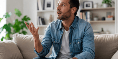 Uomo seduto sul divano di casa che parla da solo mentre riflette ad alta voce. L'immagine rappresenta il dialogo interiore, la concentrazione e i processi mentali studiati dalla psicologia.
