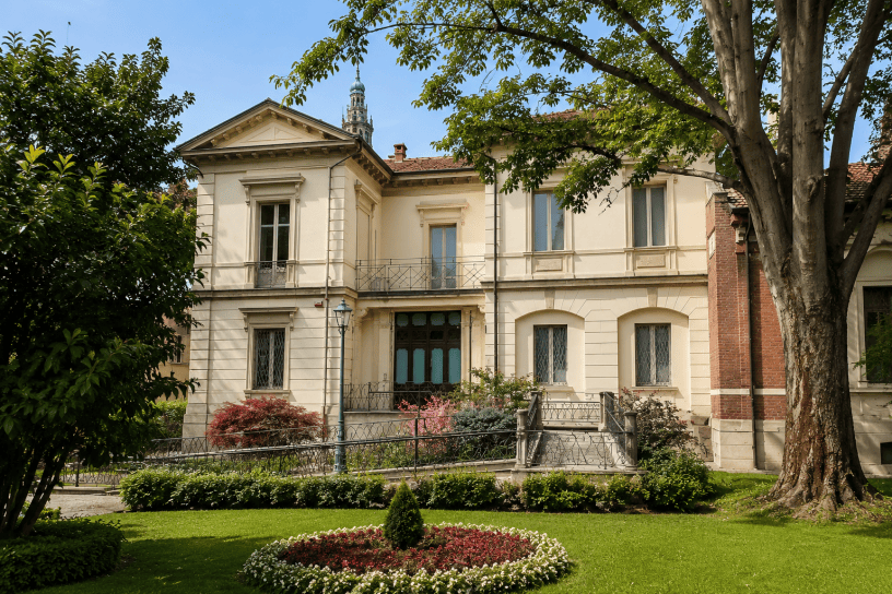 Villa Guerci ad Alessandria circondata da alberi e giardini curati, con elegante facciata storica in stile neoclassico e vista sul campanile della Cattedrale sullo sfondo.