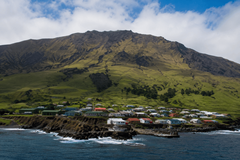 Veduta panoramica di Tristan da Cunha, l'isola abitata più isolata del mondo, con il piccolo villaggio costiero ai piedi di un'imponente montagna vulcanica affacciata sull'Oceano Atlantico.
