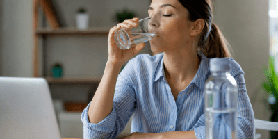 Una giovane donna seduta alla scrivania beve un bicchiere d'acqua mentre lavora al computer. Sul tavolo sono presenti una bottiglia d'acqua, un quaderno e un laptop. L'immagine rappresenta l'importanza dell'idratazione nella vita quotidiana.