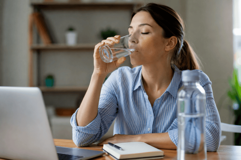 Una giovane donna seduta alla scrivania beve un bicchiere d'acqua mentre lavora al computer. Sul tavolo sono presenti una bottiglia d'acqua, un quaderno e un laptop. L'immagine rappresenta l'importanza dell'idratazione nella vita quotidiana.