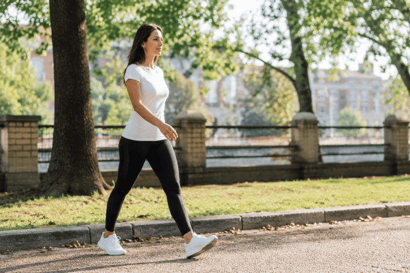 Donna che cammina con passo deciso lungo un viale alberato durante una giornata di sole, simbolo dei benefici dell'attività fisica quotidiana e di uno stile di vita sano.