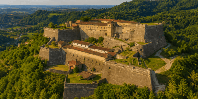 Veduta aerea panoramica di una grande fortezza storica ispirata al Forte di Gavi, arroccata su una collina boscosa e circondata dal paesaggio verde dell'Oltregiogo piemontese.