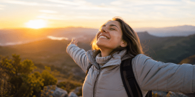Donna sorridente con le braccia aperte davanti a un panorama naturale illuminato dal sole al tramonto. L'immagine rappresenta ottimismo, benessere psicologico, speranza, resilienza e fiducia nel futuro.