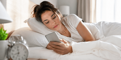 Giovane donna sdraiata nel letto al mattino mentre consulta lo smartphone appena sveglia. La luce naturale illumina la stanza e mette in evidenza una delle abitudini digitali più diffuse della vita quotidiana.