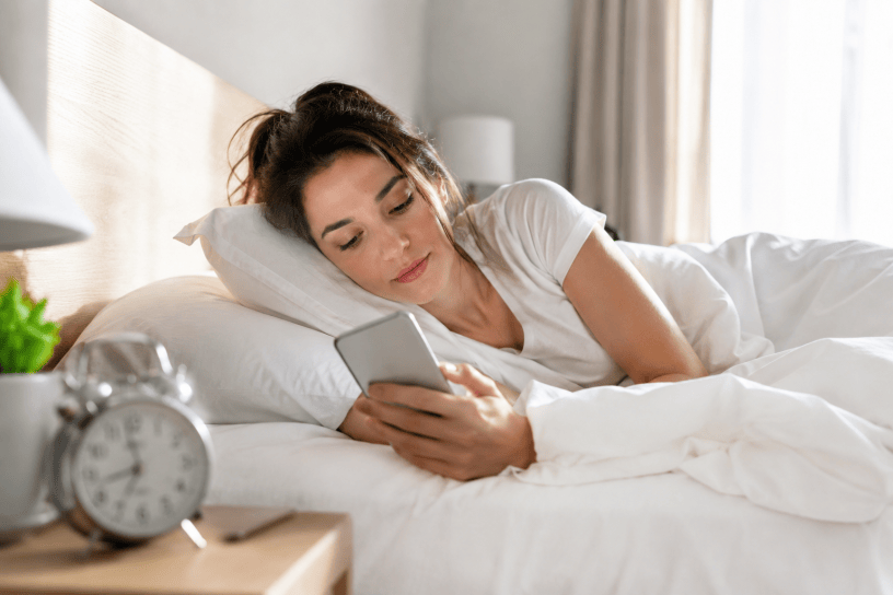 Giovane donna sdraiata nel letto al mattino mentre consulta lo smartphone appena sveglia. La luce naturale illumina la stanza e mette in evidenza una delle abitudini digitali più diffuse della vita quotidiana.