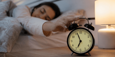 Persona che si sveglia spontaneamente pochi minuti prima della sveglia in una camera da letto illuminata da una luce soffusa. In primo piano una sveglia analogica sul comodino, simbolo dell'orologio biologico umano.