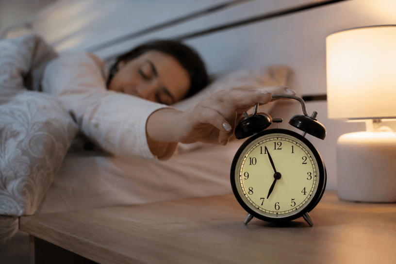 Persona che si sveglia spontaneamente pochi minuti prima della sveglia in una camera da letto illuminata da una luce soffusa. In primo piano una sveglia analogica sul comodino, simbolo dell'orologio biologico umano.