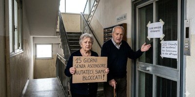 Anziani residenti in un condominio di Alessandria mostrano cartelli di protesta davanti a un ascensore fuori servizio da oltre due settimane, evidenziando il disagio per le persone con mobilità ridotta.