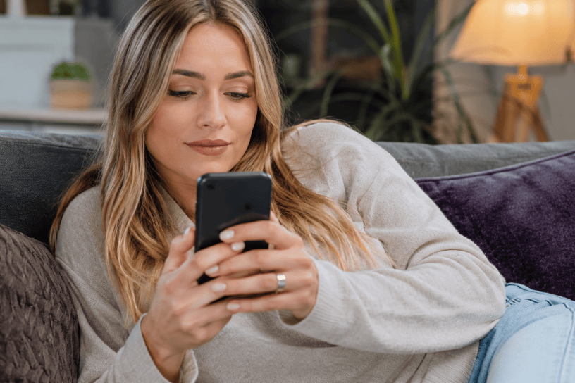 Giovane donna bionda mentre utilizza uno smartphone comodamente seduta sul divano di casa, simbolo dell'uso quotidiano della tecnologia e dei dispositivi mobili.