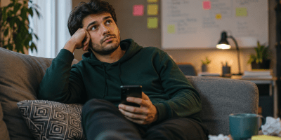 Uomo seduto sul divano mentre osserva il vuoto con lo smartphone in mano, immerso nei propri pensieri e nella tendenza a rimandare decisioni e impegni importanti. L'immagine rappresenta la procrastinazione e la cosiddetta sindrome del "ci penso domani".