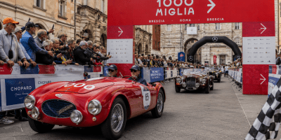Una fila di splendide auto d'epoca percorre una strada italiana panoramica durante la 1000 Miglia. Le vetture storiche sfilano tra il pubblico e i paesaggi caratteristici del Paese, rievocando il fascino dell'automobilismo del Novecento.