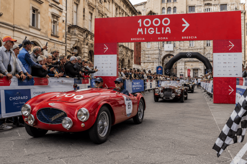 Una fila di splendide auto d'epoca percorre una strada italiana panoramica durante la 1000 Miglia. Le vetture storiche sfilano tra il pubblico e i paesaggi caratteristici del Paese, rievocando il fascino dell'automobilismo del Novecento.