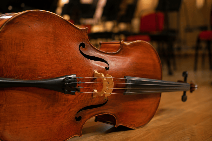 Primo piano di un violoncello in legno pregiato fotografato su un palcoscenico da concerto, con illuminazione calda e sfondo sfocato di una sala musicale.