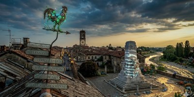 Una panoramica fotorealistica di una storica città italiana al tramonto. In primo piano, su un tetto, spicca un segnavento a forma di gallo, reso con uno stile biologico e bioluminescente, che sorregge una serie di cartelli in metallo antico con iscrizioni in italiano: "TESTO ALTERNATIVO", "ALTERNATLVO", "TESTO DEL TITOLO", "FRASE PER IMMAGINI", "YA DIDASCALIA" e "E TESTO DELLA DESCRIZIONE". Dietro, un labirinto di tetti in terracotta conduce a una torre campanaria con orologio. Sulla destra, una piazza pavimentata accoglie un imponente monumento di marmo, avvolto da simboli e testi proiettati. Oltre, si estende il letto asciutto di un fiume e colline alberate sotto un cielo nuvoloso e drammatico.