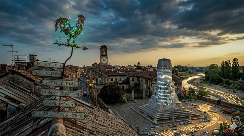 Una panoramica fotorealistica di una storica città italiana al tramonto. In primo piano, su un tetto, spicca un segnavento a forma di gallo, reso con uno stile biologico e bioluminescente, che sorregge una serie di cartelli in metallo antico con iscrizioni in italiano: "TESTO ALTERNATIVO", "ALTERNATLVO", "TESTO DEL TITOLO", "FRASE PER IMMAGINI", "YA DIDASCALIA" e "E TESTO DELLA DESCRIZIONE". Dietro, un labirinto di tetti in terracotta conduce a una torre campanaria con orologio. Sulla destra, una piazza pavimentata accoglie un imponente monumento di marmo, avvolto da simboli e testi proiettati. Oltre, si estende il letto asciutto di un fiume e colline alberate sotto un cielo nuvoloso e drammatico.