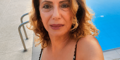Ritratto di donna sorridente in primo piano accanto a una piscina all'aperto durante una giornata estiva, con capelli mossi e abito elegante dai dettagli luminosi.