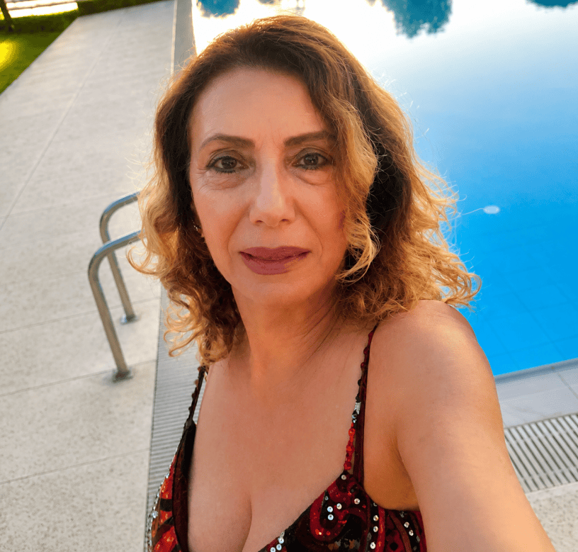 Ritratto di donna sorridente in primo piano accanto a una piscina all'aperto durante una giornata estiva, con capelli mossi e abito elegante dai dettagli luminosi.