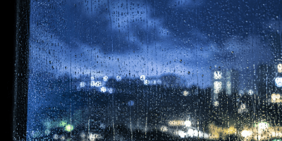 Gocce di pioggia scorrono lentamente su una finestra illuminata dalla luce notturna, mentre sullo sfondo sfocato si intravedono luci urbane e un cielo carico di nuvole estive.