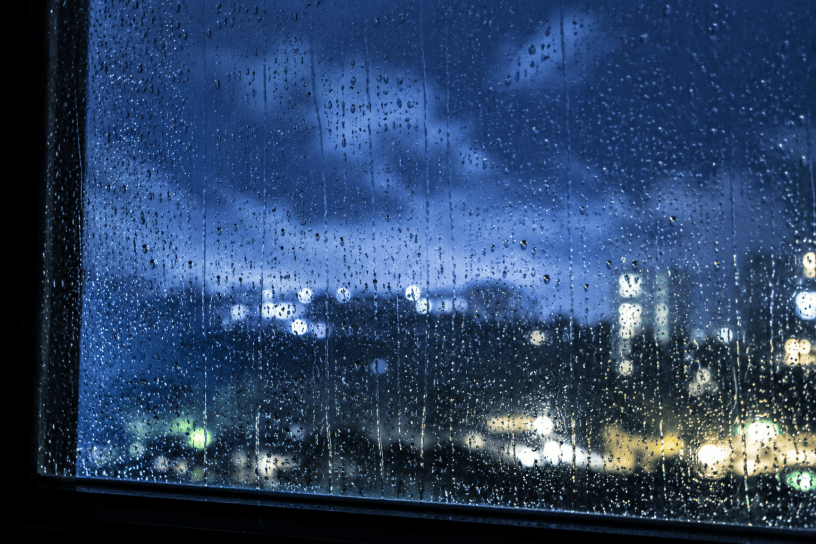 Gocce di pioggia scorrono lentamente su una finestra illuminata dalla luce notturna, mentre sullo sfondo sfocato si intravedono luci urbane e un cielo carico di nuvole estive.