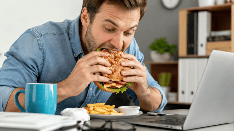 Uomo mangia rapidamente un hamburger davanti al computer durante una pausa di lavoro, simbolo delle cattive abitudini alimentari e dei possibili effetti sulla salute.