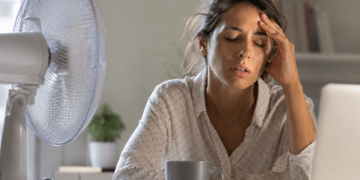 Giovane donna visibilmente affaticata davanti al computer durante una giornata calda, con ventilatore acceso e segni di stanchezza dovuti alle alte temperature.