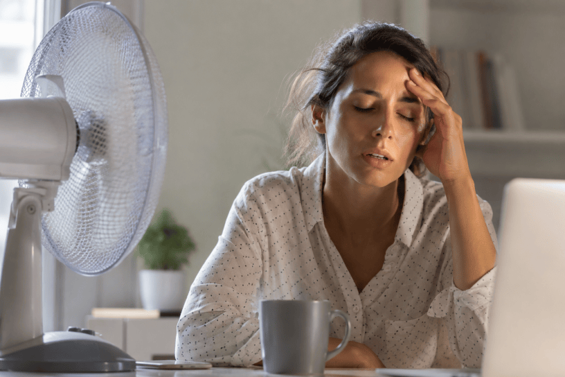 Giovane donna visibilmente affaticata davanti al computer durante una giornata calda, con ventilatore acceso e segni di stanchezza dovuti alle alte temperature.