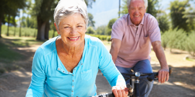 Coppia di anziani sorridenti in bicicletta lungo un sentiero immerso nel verde durante una giornata luminosa, simbolo di salute, benessere e longevità attiva.