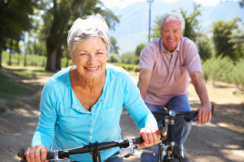 Coppia di anziani sorridenti in bicicletta lungo un sentiero immerso nel verde durante una giornata luminosa, simbolo di salute, benessere e longevità attiva.