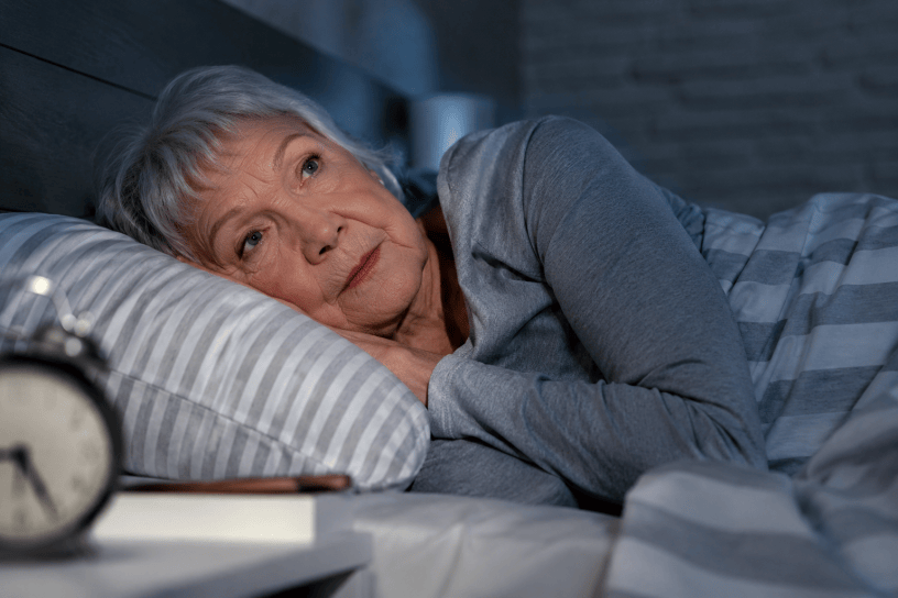 Donna anziana sdraiata nel letto durante la notte mentre osserva il soffitto senza riuscire a dormire. L'immagine rappresenta i disturbi del sonno che possono comparire dopo i 60 anni.