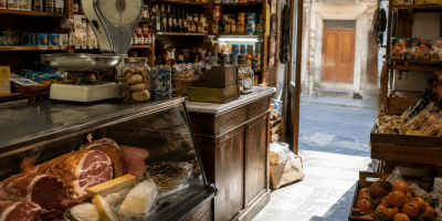 Interno di una tradizionale bottega di paese con banco dei salumi e dei formaggi, scaffali pieni di conserve, frutta fresca e atmosfera calda che richiama i negozi di una volta.