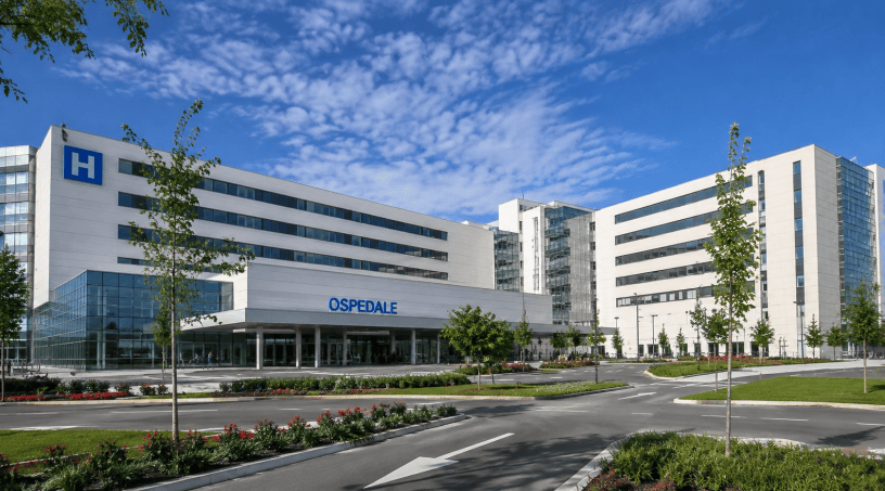 Grande ospedale moderno di nuova generazione con facciate in vetro e acciaio, ampi spazi verdi, viabilità dedicata e ingresso principale progettato secondo i più avanzati standard sanitari.