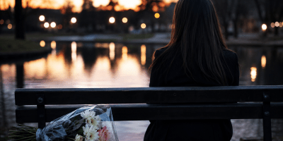 Donna seduta sola su una panchina al tramonto davanti a uno specchio d'acqua, con un mazzo di fiori accanto, in un'atmosfera malinconica e contemplativa.