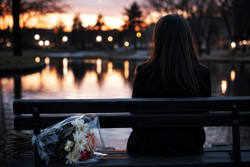 Donna seduta sola su una panchina al tramonto davanti a uno specchio d'acqua, con un mazzo di fiori accanto, in un'atmosfera malinconica e contemplativa.