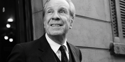 Ritratto fotografico in bianco e nero ispirato a Jorge Luis Borges, uomo anziano in abito elegante che sorride leggermente mentre osserva verso l’alto, in un contesto urbano dall’atmosfera letteraria e senza tempo.