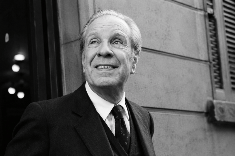 Ritratto fotografico in bianco e nero ispirato a Jorge Luis Borges, uomo anziano in abito elegante che sorride leggermente mentre osserva verso l’alto, in un contesto urbano dall’atmosfera letteraria e senza tempo.