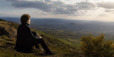 Giacomo Leopardi osserva un vasto paesaggio collinare italiano, immerso nella natura e nella contemplazione dell’infinito, in una scena fotografica ispirata al suo pensiero sul rapporto tra uomo e ambiente.