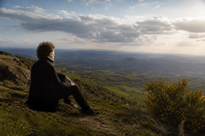 Giacomo Leopardi osserva un vasto paesaggio collinare italiano, immerso nella natura e nella contemplazione dell’infinito, in una scena fotografica ispirata al suo pensiero sul rapporto tra uomo e ambiente.