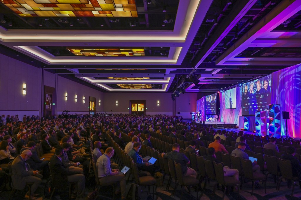 A large audience seated in a conference hall, with a speaker on stage and multiple screens displaying graphics and speakers' images.