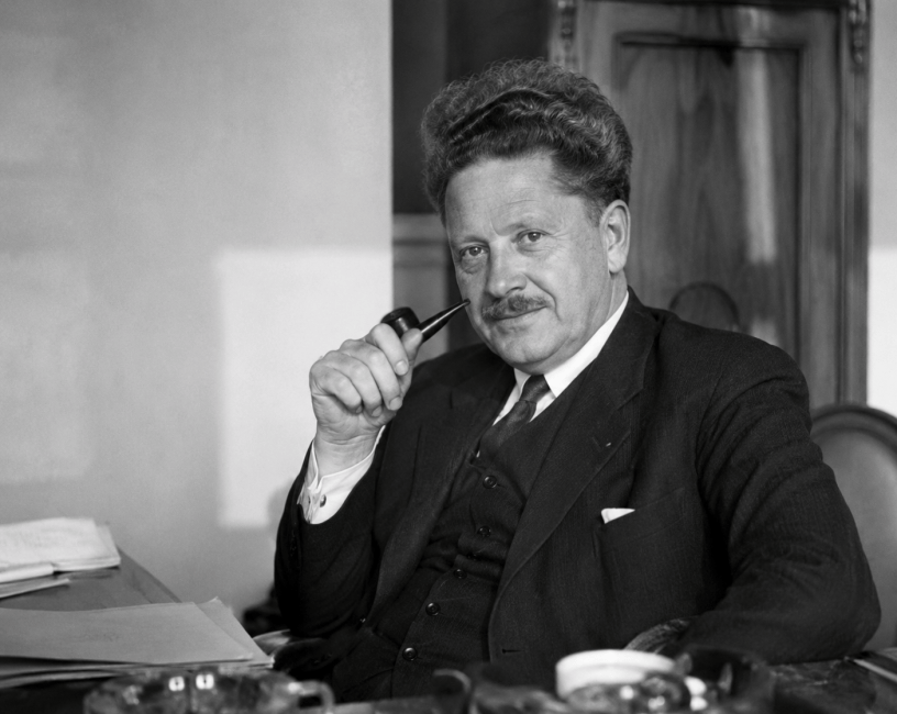 Ritratto fotografico in bianco e nero di Nazım Hikmet seduto alla scrivania con una pipa in mano. Il celebre poeta turco appare pensieroso e riflessivo in un ambiente di lavoro d'epoca.