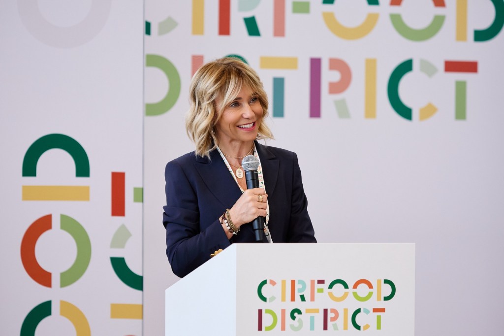 A woman with blonde hair speaking into a microphone at an event, with a backdrop featuring colorful letters that read 'CIRFOOD DISTRICT'.