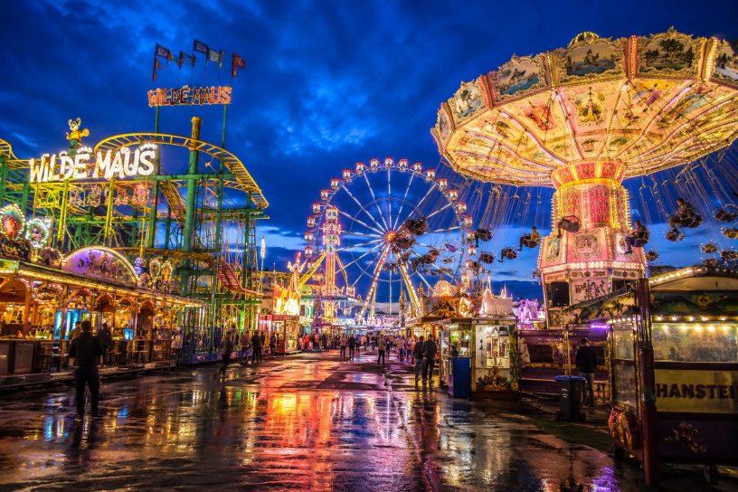 Grande Luna Park illuminato di sera con ruota panoramica, giostre colorate e luci riflesse sul pavimento bagnato, in un'atmosfera suggestiva e ricca di emozioni.
