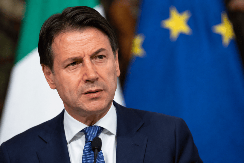 Ritratto realistico di Giuseppe Conte durante una conferenza stampa istituzionale. L'ex Presidente del Consiglio appare con espressione seria e concentrata mentre affronta temi di attualità politica nazionale.