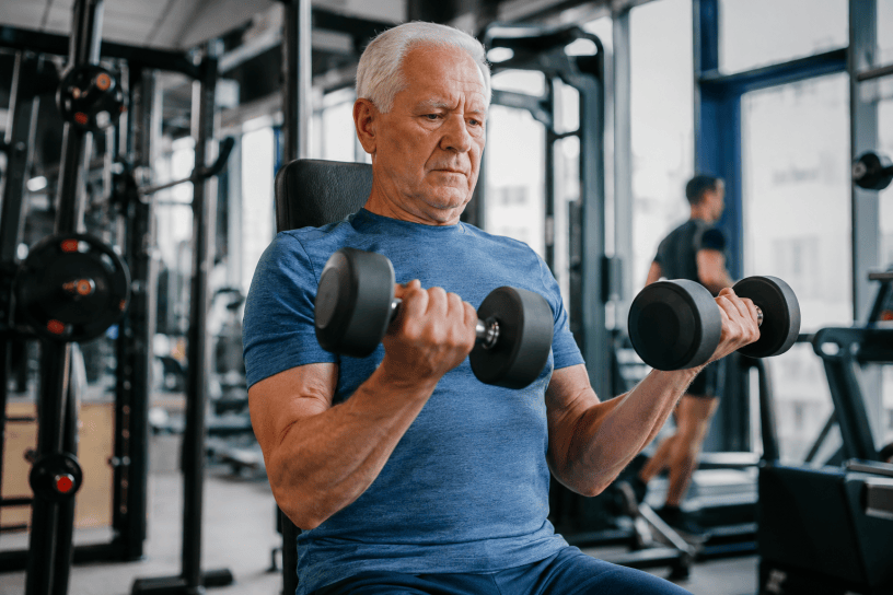 Anziano che si allena con manubri in palestra per mantenere forza muscolare e contrastare la sarcopenia, in un ambiente moderno e luminoso.